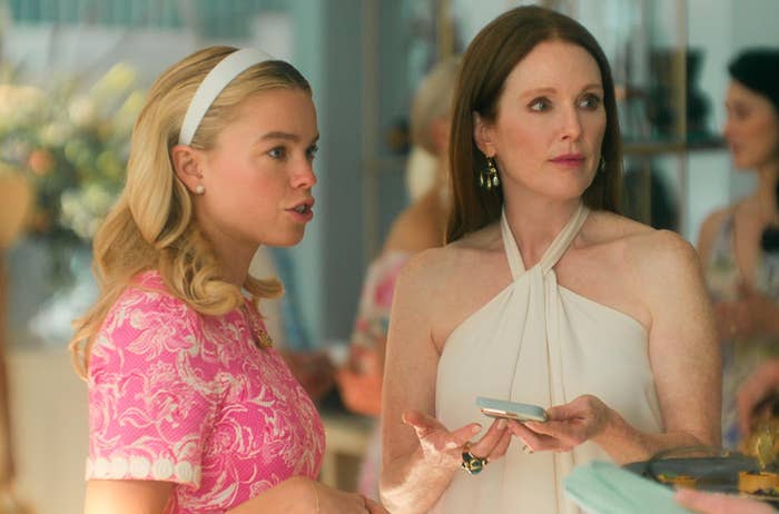 Two women in conversation at an event; one wears a pink patterned dress, the other a white halter dress, both holding objects in their hands