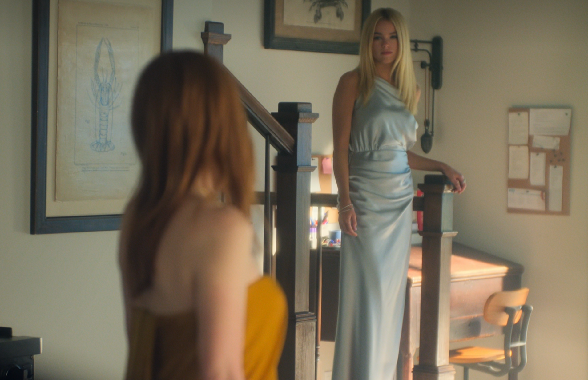 Two women stand in a room; one in a sleek dress on stairs, the other in an off-shoulder gown, facing each other. Scene from a TV show