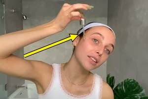 Woman applying skincare serum to her face using a dropper, standing in a bathroom with a plant visible in the background