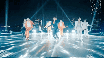 Seven performers dance energetically under spotlights on a rooftop with city views, wearing stylish suits in varied designs