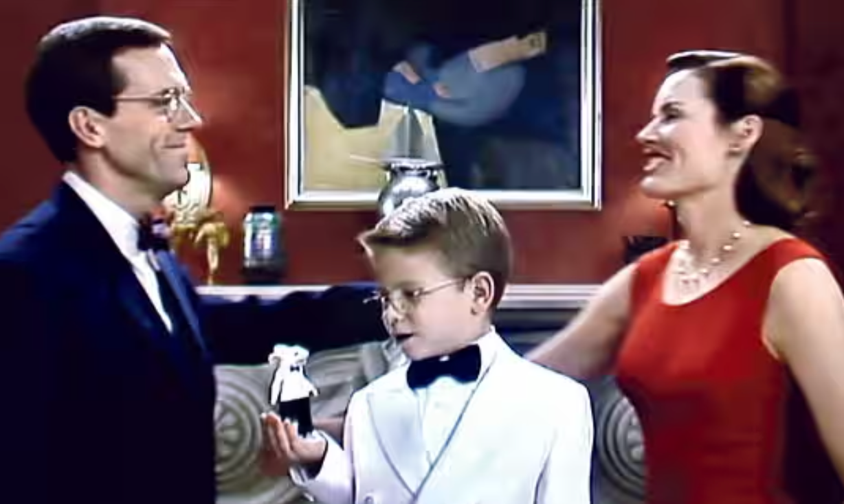 A young boy in a white tuxedo stands between a smiling man in a suit and a woman in an elegant dress at a formal event