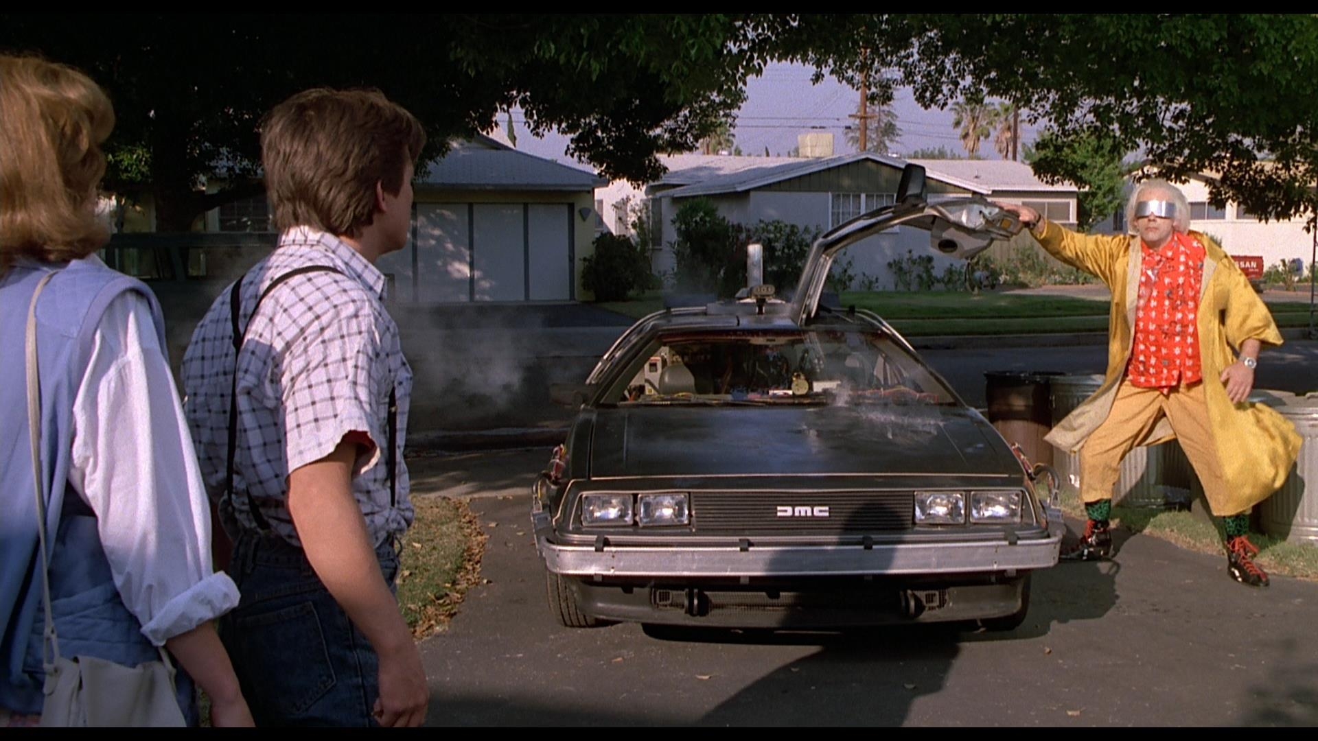 Two people look at a man in an eccentric outfit excitedly pointing at a smoking DeLorean car with open gullwing doors