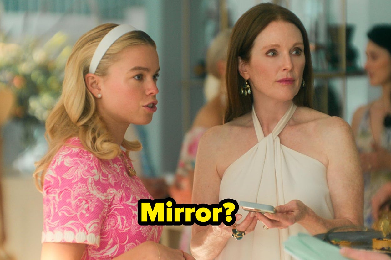 Two women in conversation at an event; one wears a pink patterned dress, the other a white halter dress, both holding objects in their hands