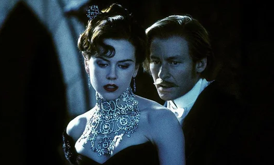 Two actors in formal attire; one wears a lavish necklace and strapless gown, the other a suit. They share an intense, dramatic moment