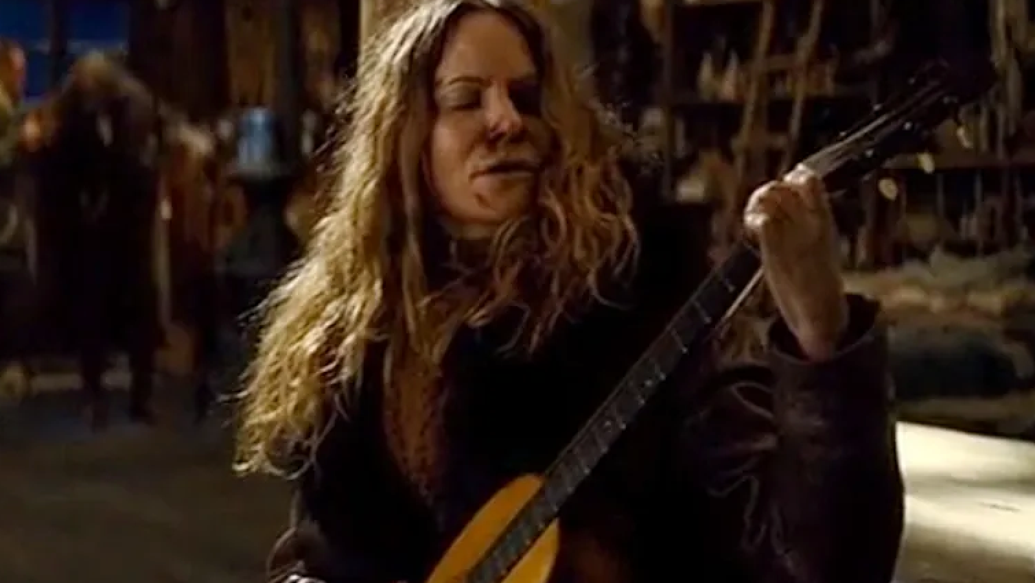 A person with long hair plays a guitar in a rustic indoor setting filled with various objects