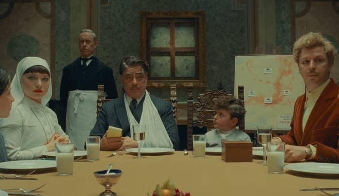 Five people sit at a formal dining table, all wearing elegant attire. A map is visible in the background