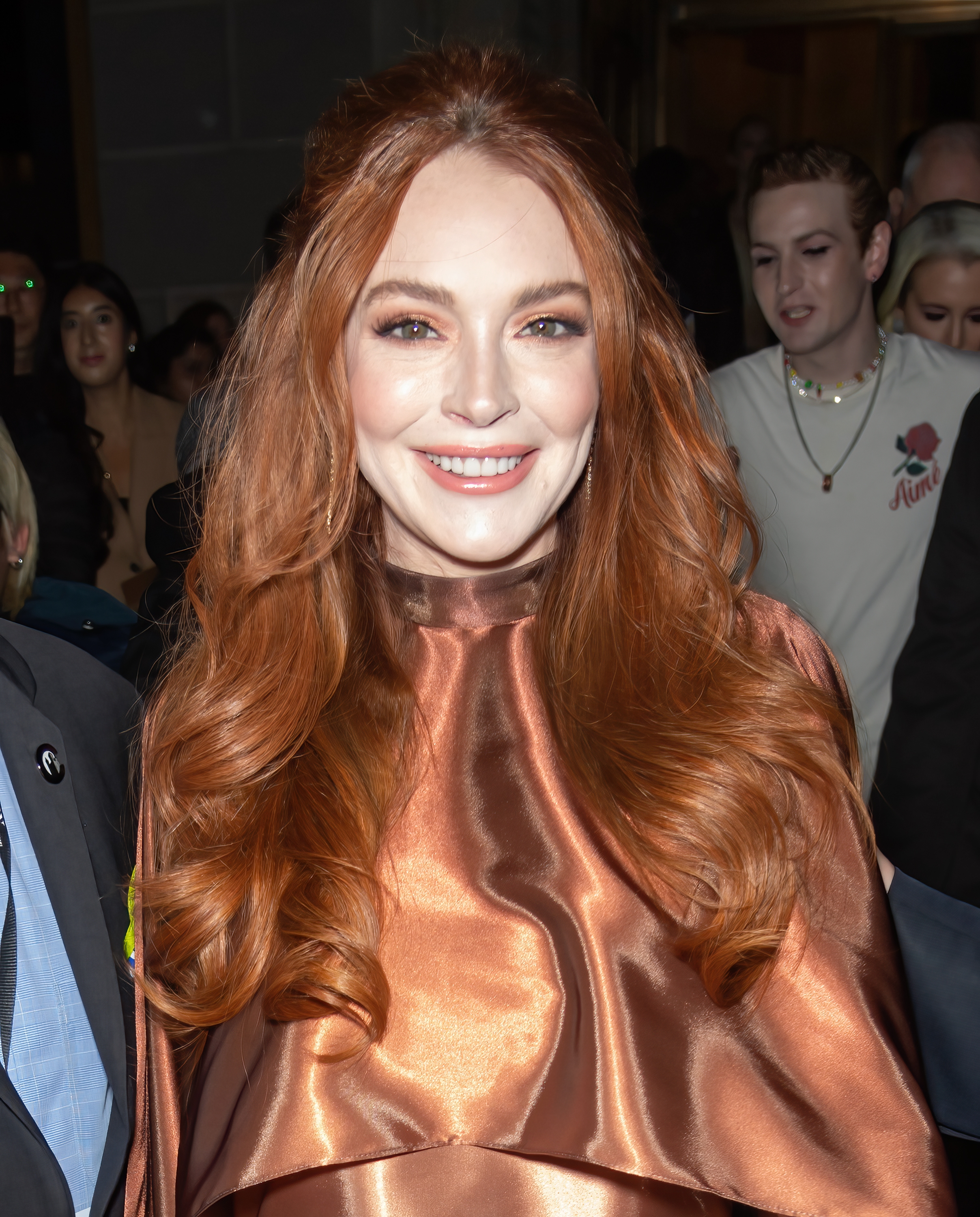 Person with styled long hair wearing a shiny, elegant top, smiling at a public event. Crowd in the background