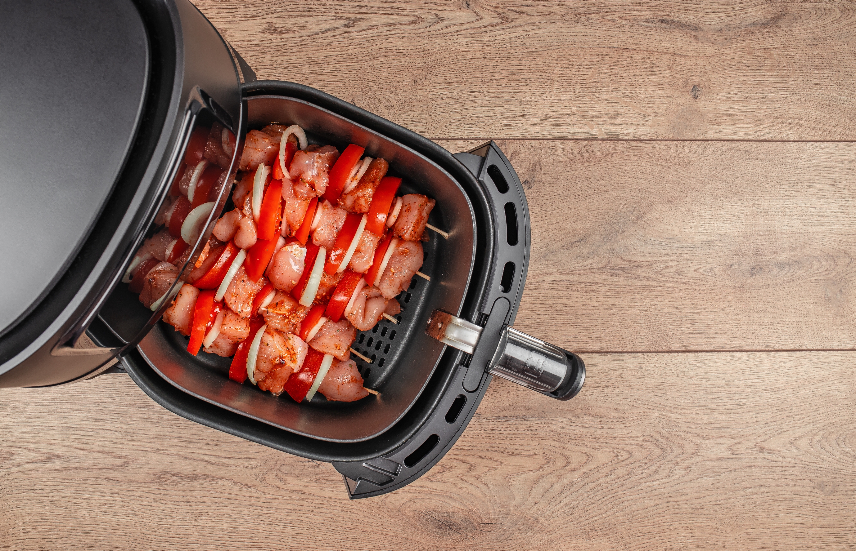 Air fryer with skewers of seasoned chicken and sliced vegetables inside, ready for cooking on a wooden countertop