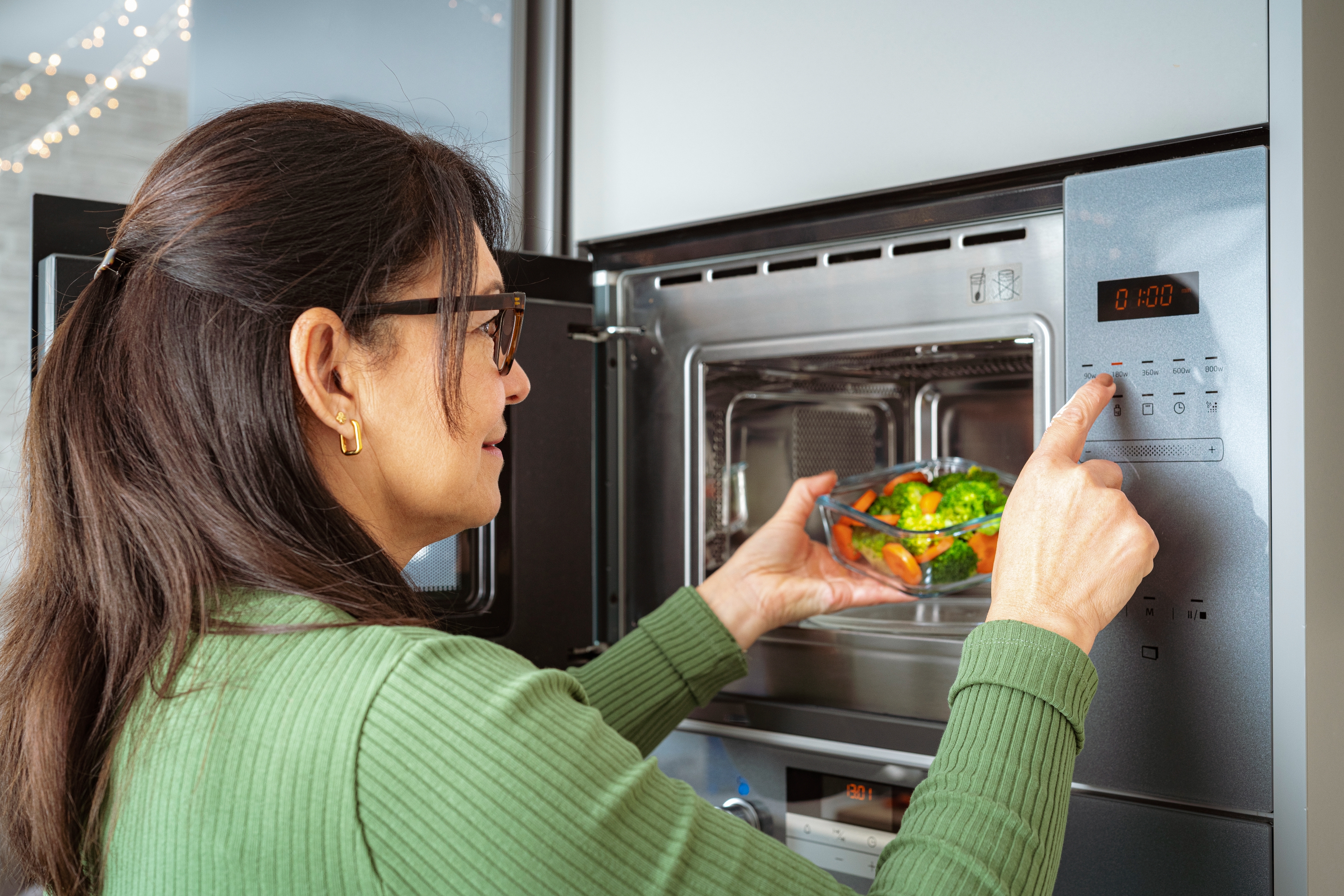 Woman in glasses microwaves a dish with vegetables, pressing buttons on the appliance