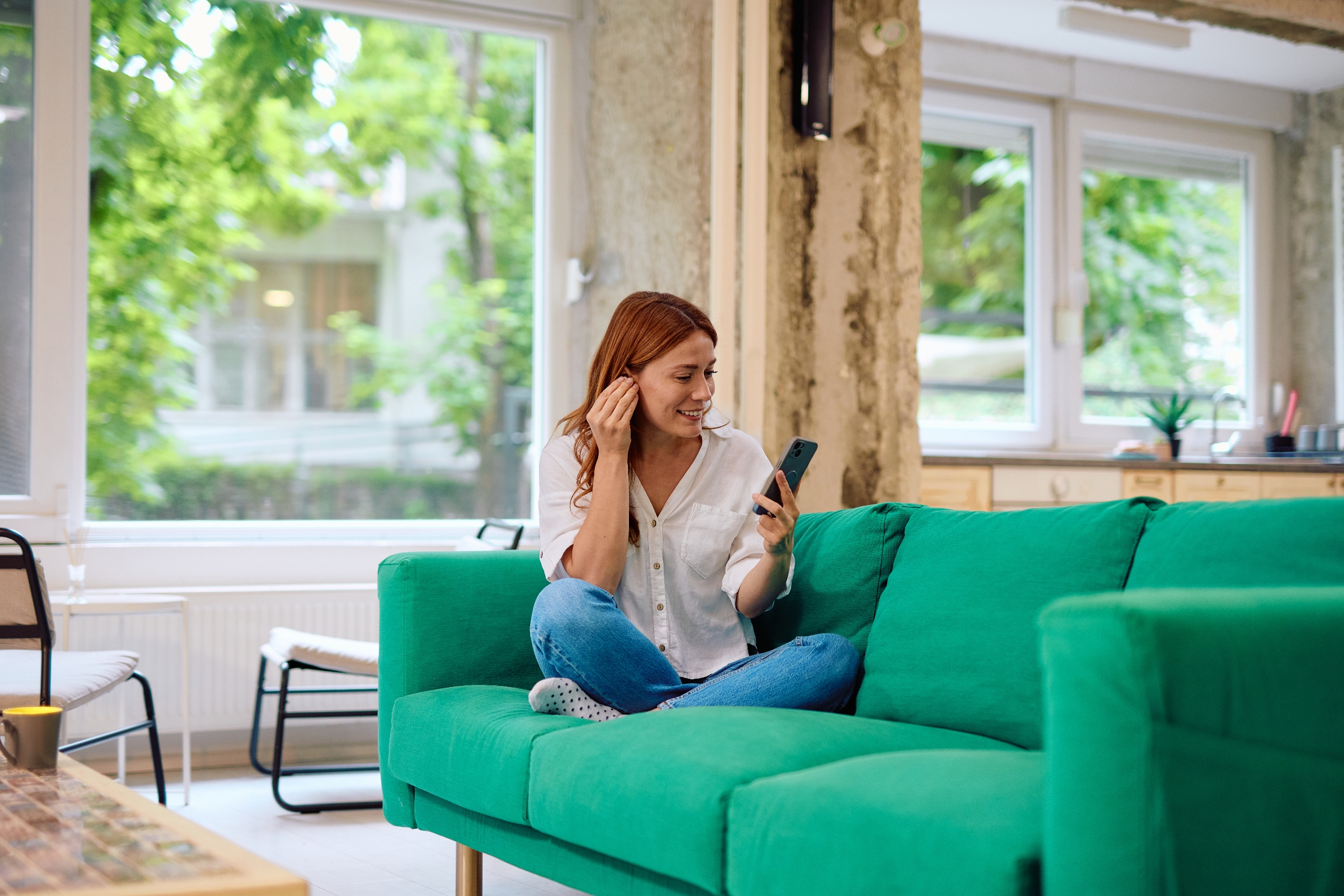 Person with long hair sits cross-legged on a couch, smiling at a phone in a cozy living room with large windows