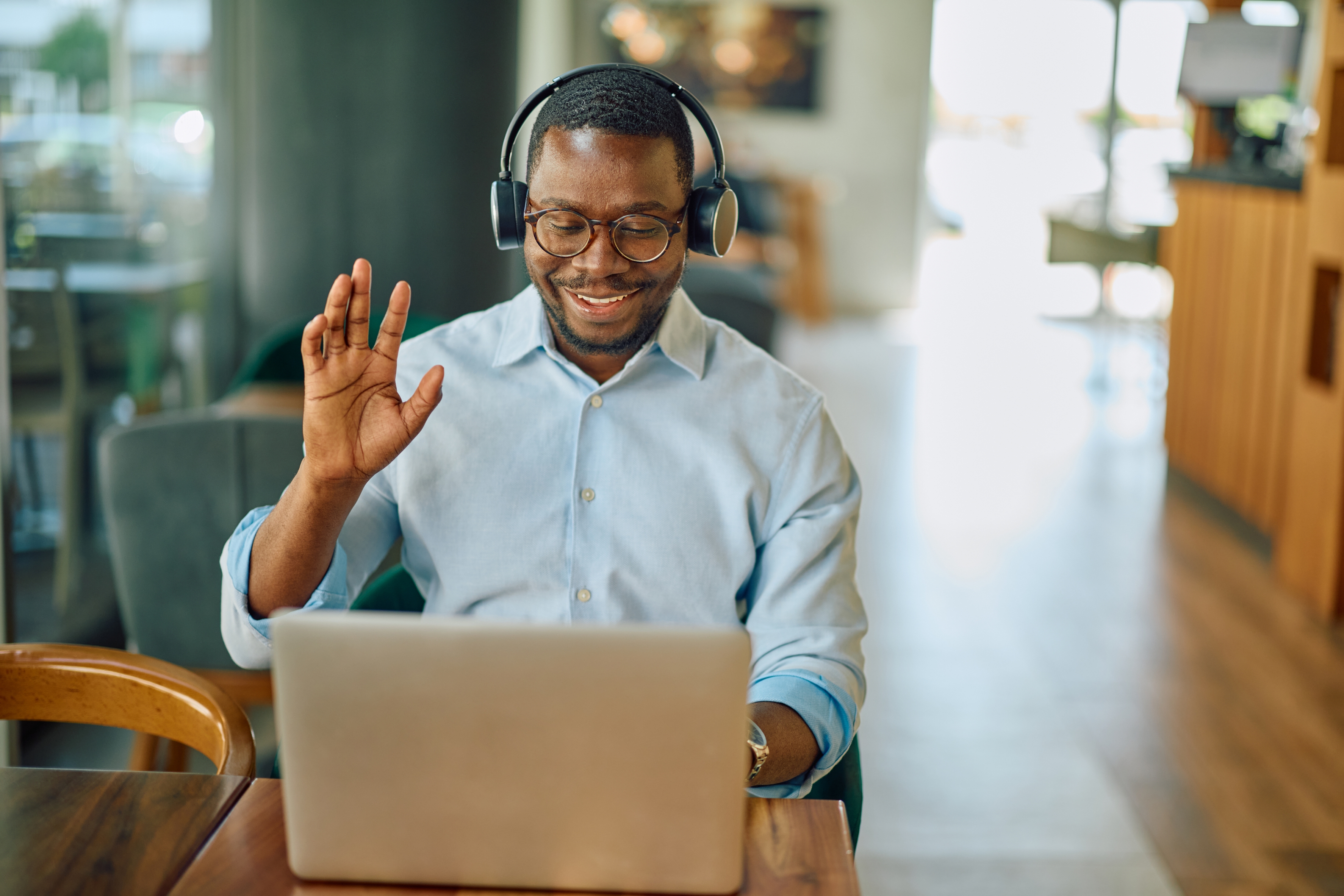 Man in headphones waves at laptop during video call, sitting indoors at a wooden table