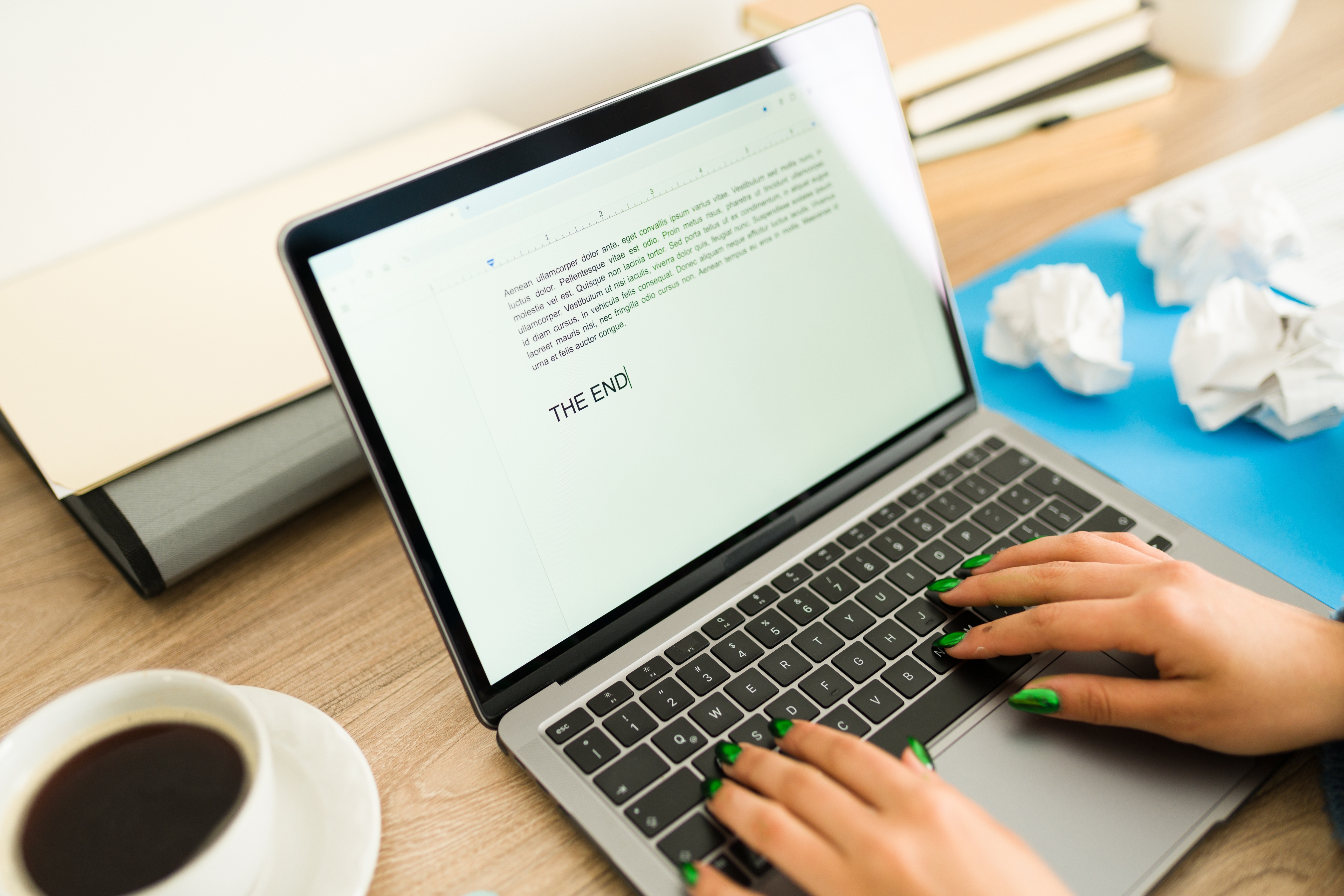 Person typing on a laptop at a desk. Nearby are crumpled papers, a cup of coffee, and a folder. Text on the screen ends with "THE END."