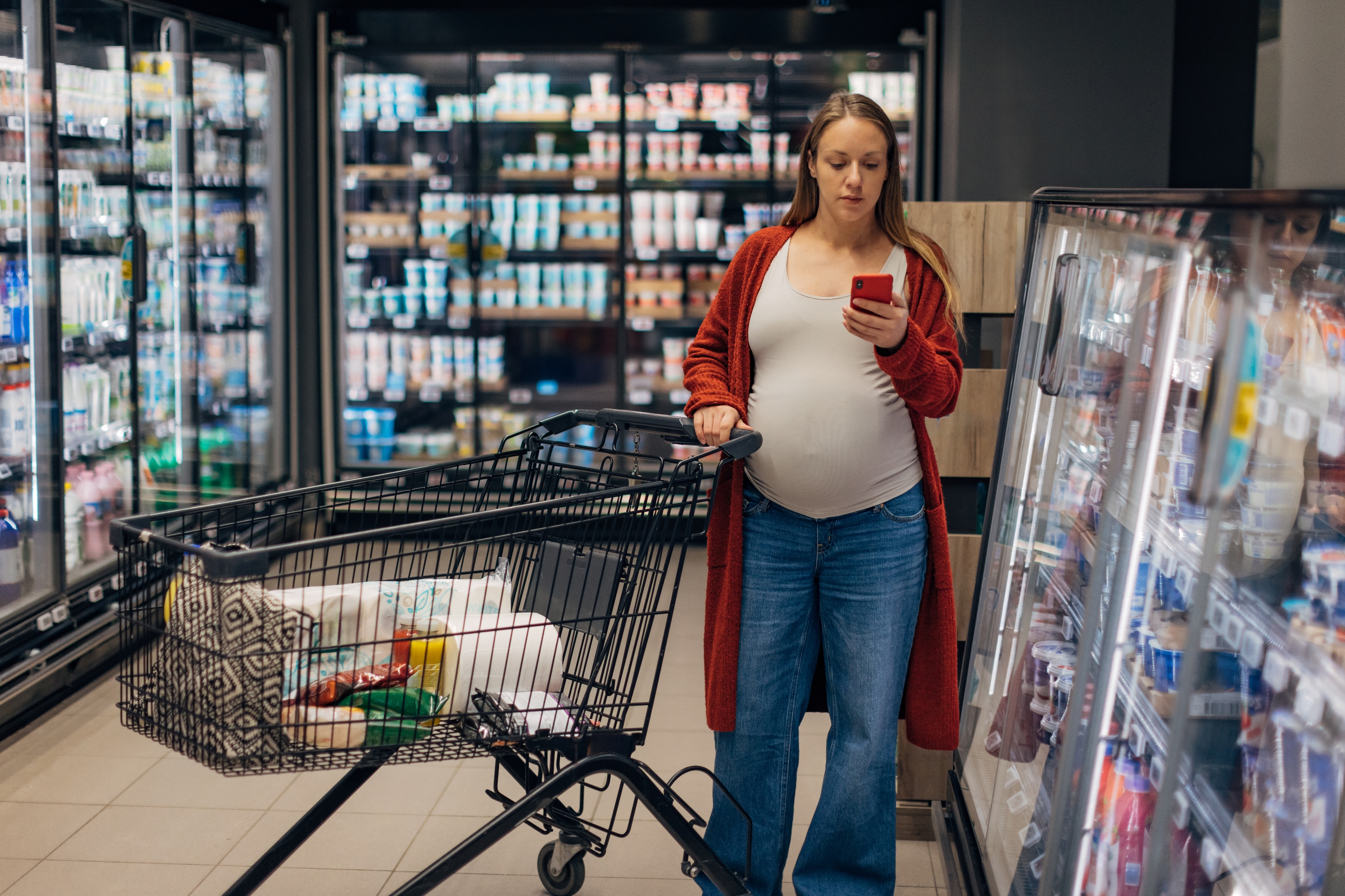 Pregnant person in a grocery store aisle, wearing casual jeans and a long cardigan, looks at their phone while holding a shopping cart