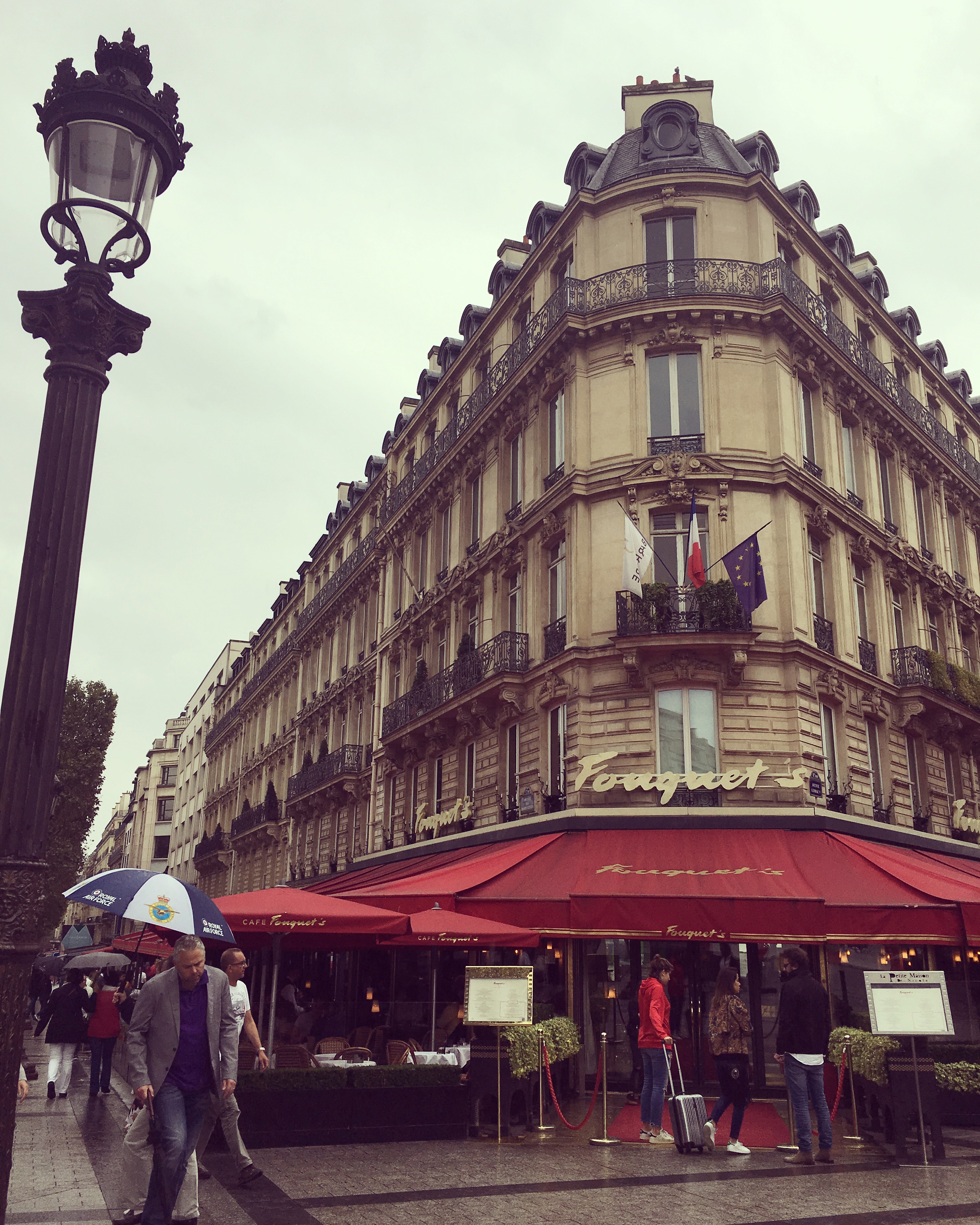 People walking by Fouquet's restaurant on a rainy day in Paris, with a streetlamp and historic building in view