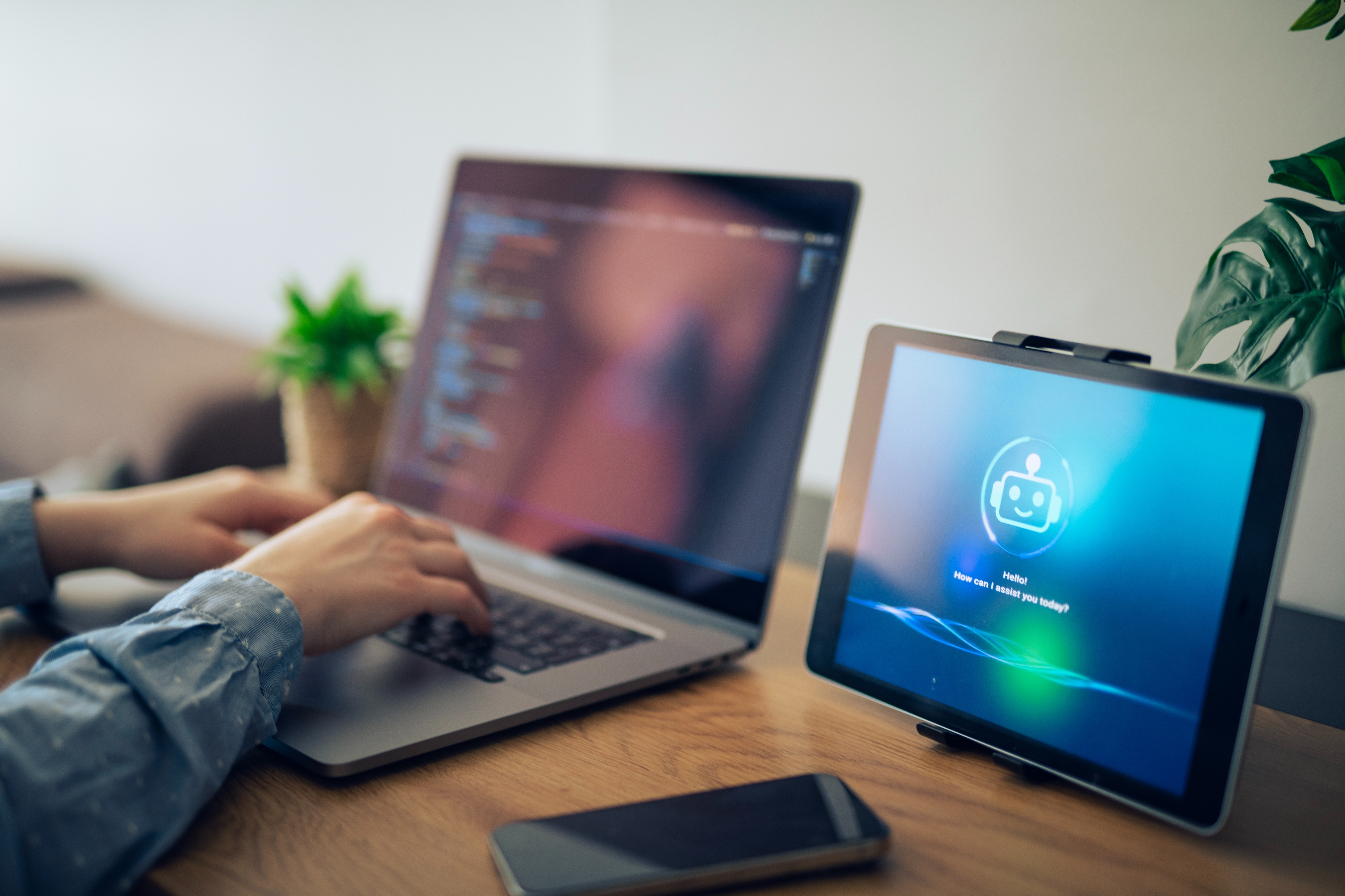 Person typing on a laptop at a desk with a tablet displaying a smiley face, symbolizing AI, next to them. A smartphone and plant are also on the desk