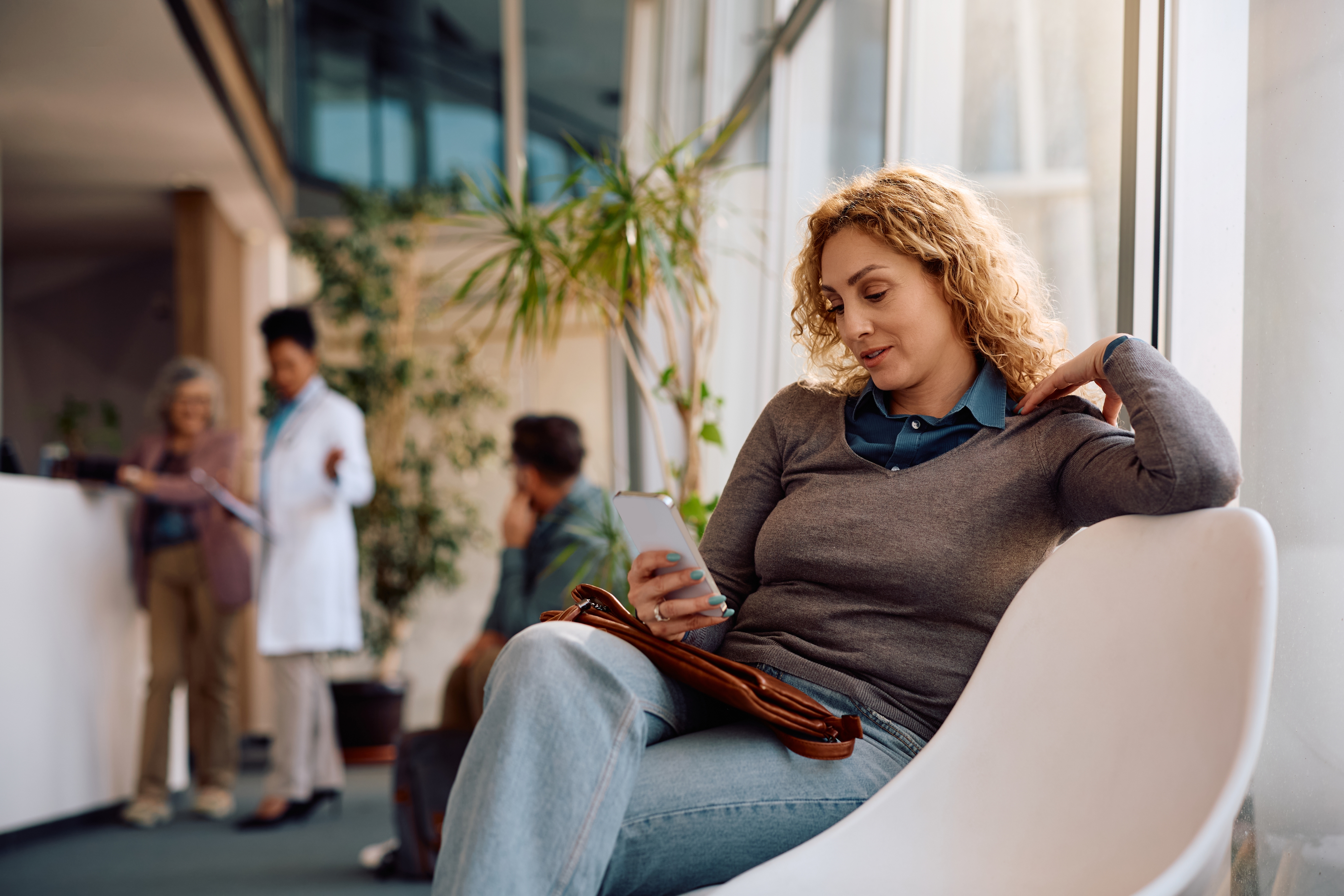 Person sitting in a waiting area, looking at their phone, with others and a doctor in the background