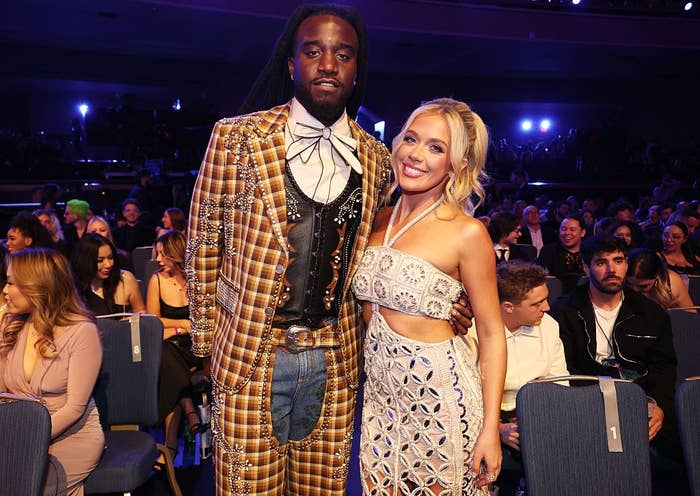 Two people on stage. One wears a patterned suit with a bowtie; the other wears a patterned halter dress. Audience in the background