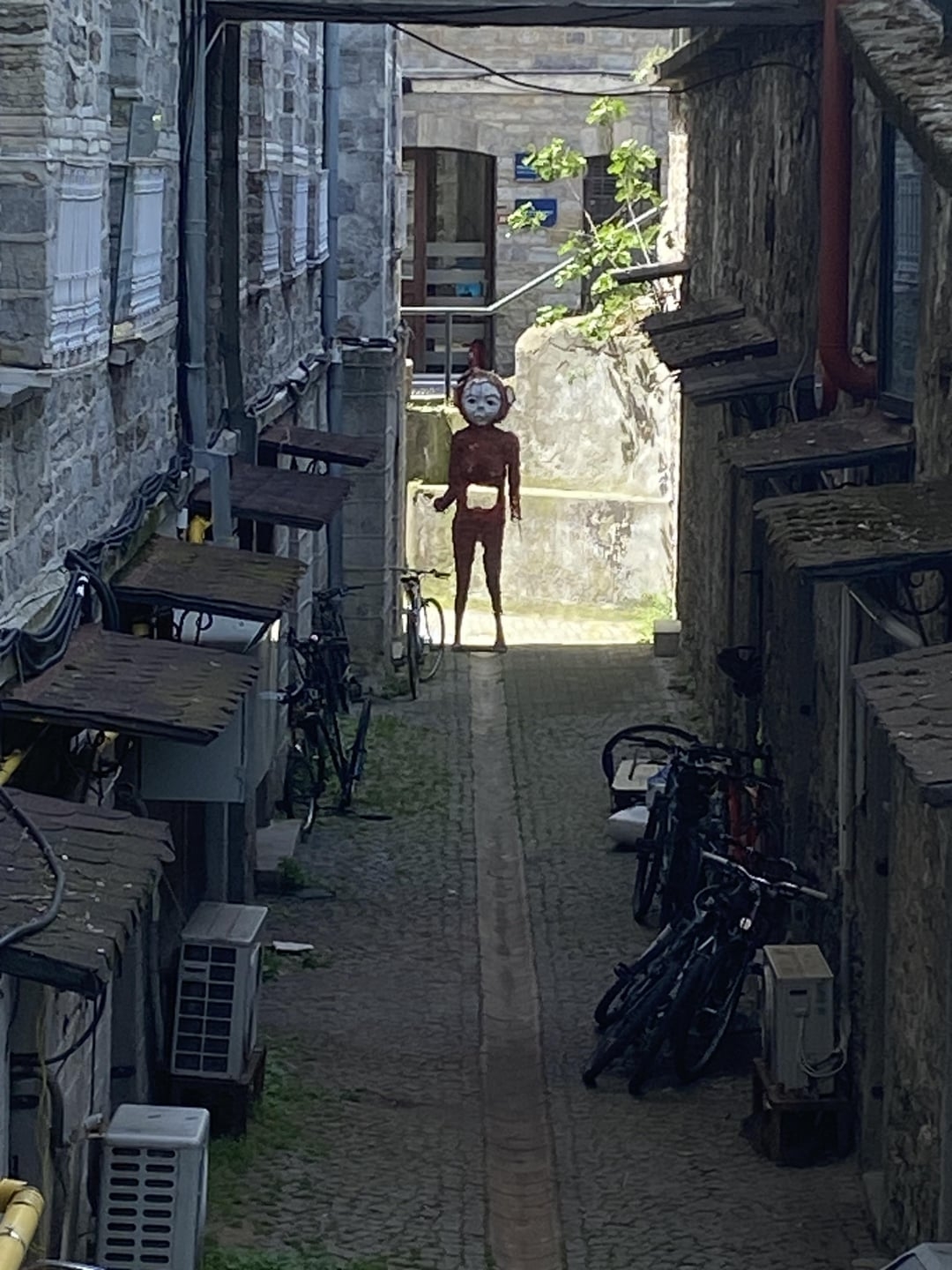 A person with a clock for a face stands at the end of a narrow alley lined with bicycles and buildings