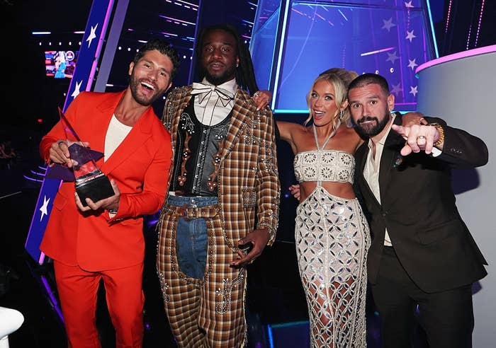 Four people at an event. One holds a trophy. Attire includes a bright suit, plaid suit, tailored dress with cut-out details, and dark suit. All are smiling