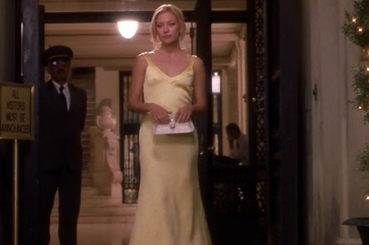 Person in a sleek, elegant gown holds a clutch while standing in a lit entryway. A uniformed doorman is nearby