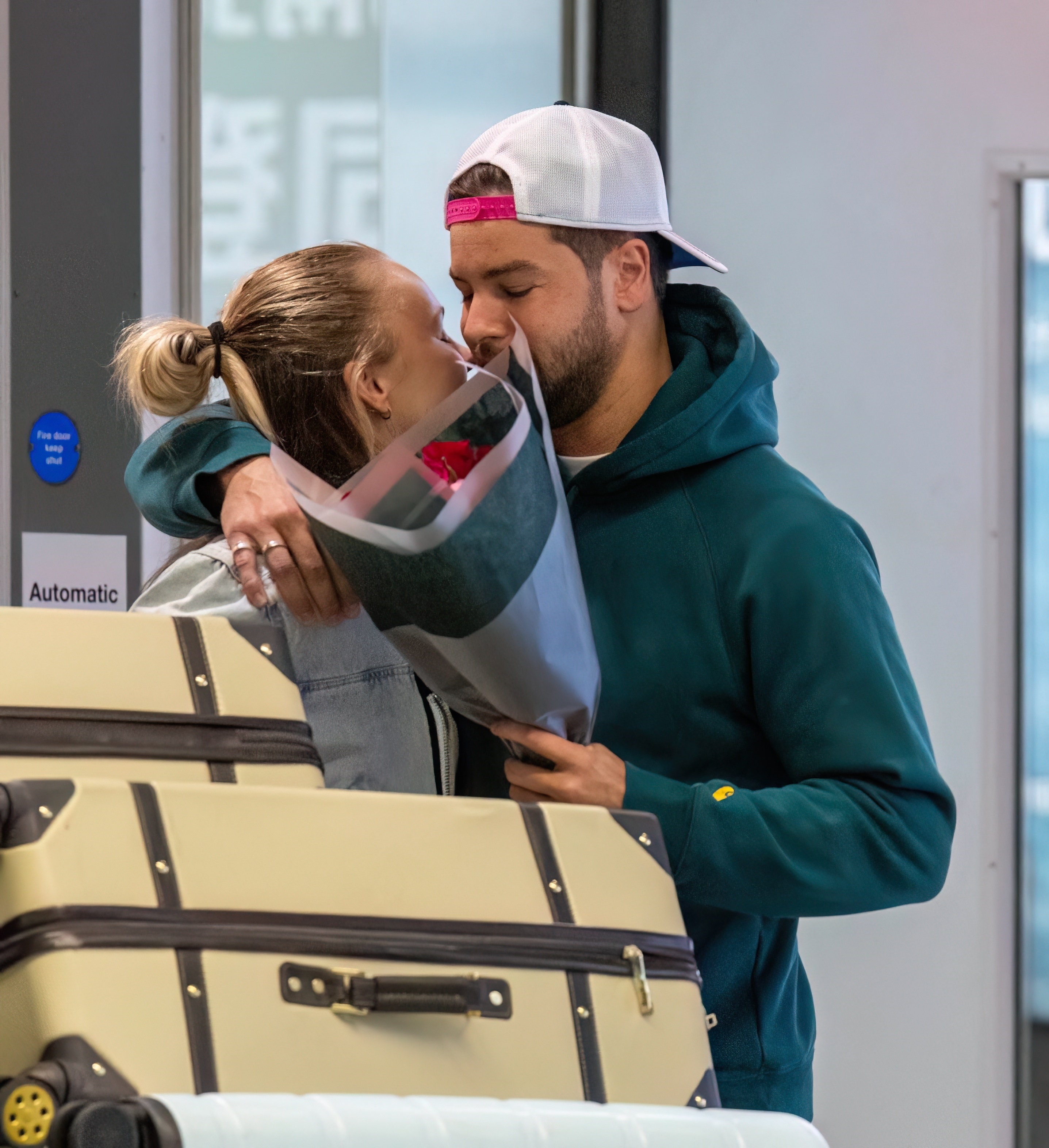 A couple hugging and kissing at an airport, holding a bouquet of roses, with suitcases nearby