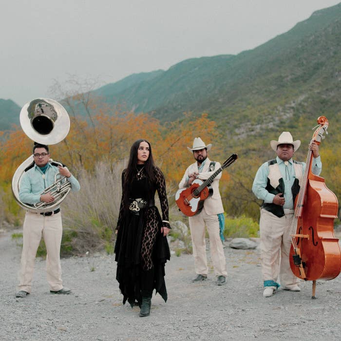 Grupo musical en exterior montañoso. Mujer en vestido negro con cinturón decorado, rodeada de músicos con instrumentos de viento y cuerda, con sombreros