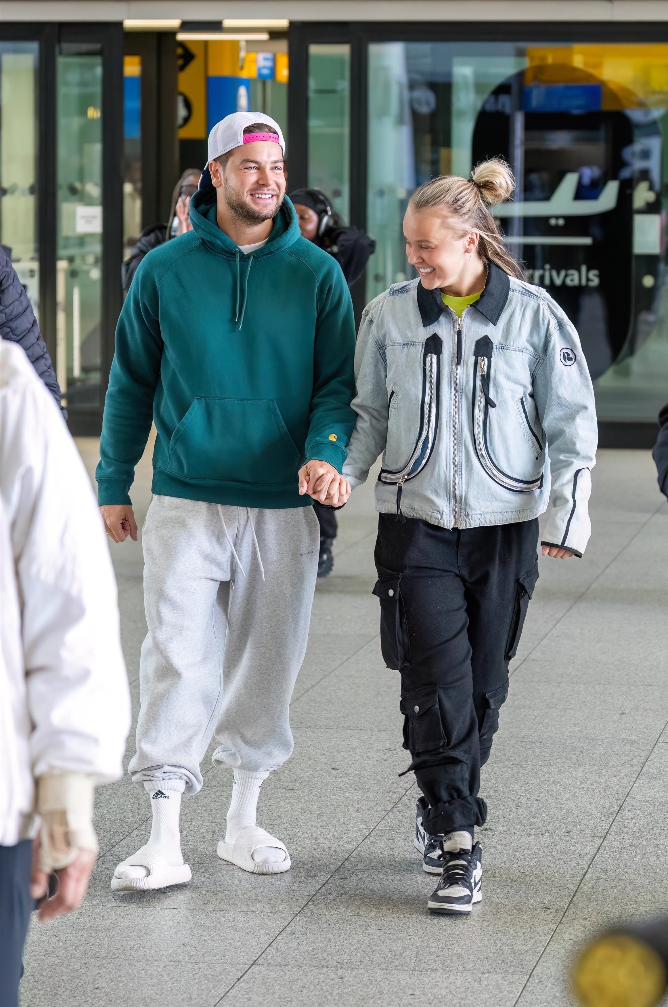 Two people smiling, casually dressed, holding hands at an airport. One wears a hoodie and sweatpants; the other wears a jacket and cargo pants