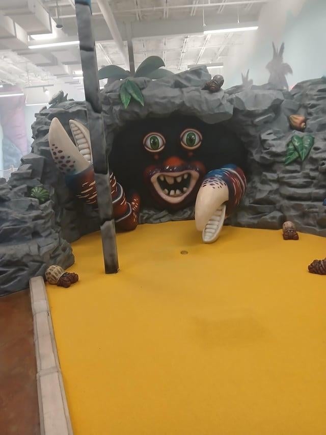 Giant crab creature with wide eyes and big claws emerging from a cave in a themed indoor mini-golf setting