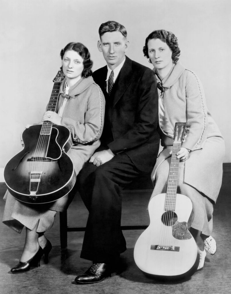 Three individuals seated; two women holding guitars, dressed in vintage 1920s-style clothing, alongside a man in a suit and tie