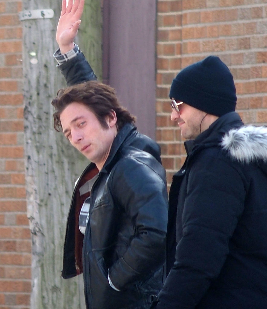 Two individuals walking and talking outside on a sidewalk. The person on the left raises a hand, wearing a leather jacket. The other wears a beanie and sunglasses