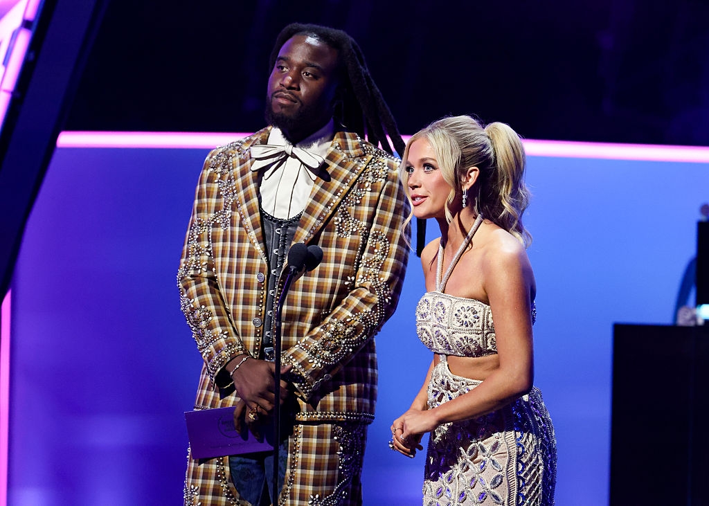 Two people stand on stage. The man wears a plaid coat with metallic details; the woman is in a strapless, embellished gown. They are presenting an award
