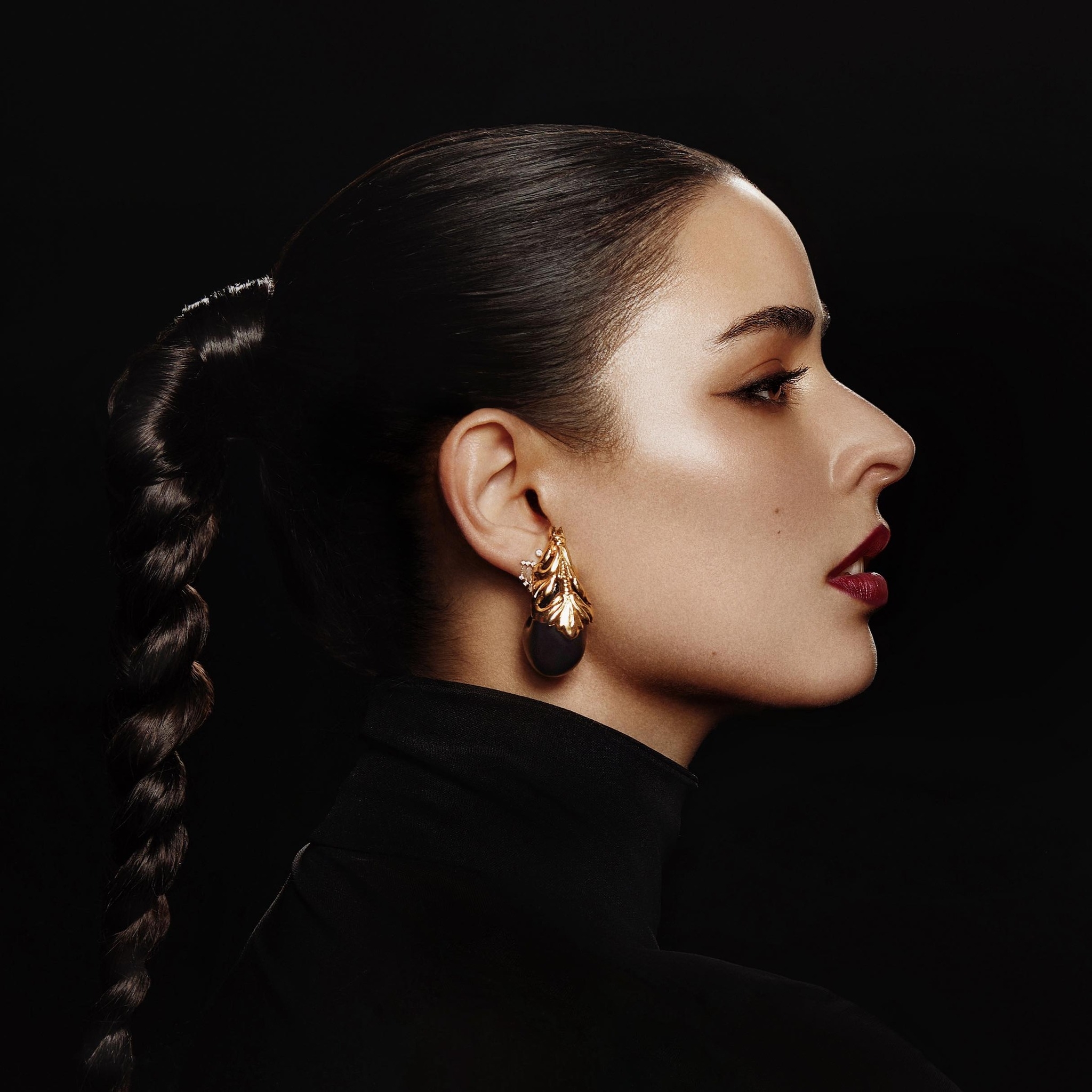 Mujer con un peinado recogido y un pendiente dorado en forma de hoja, perfilada sobre un fondo oscuro