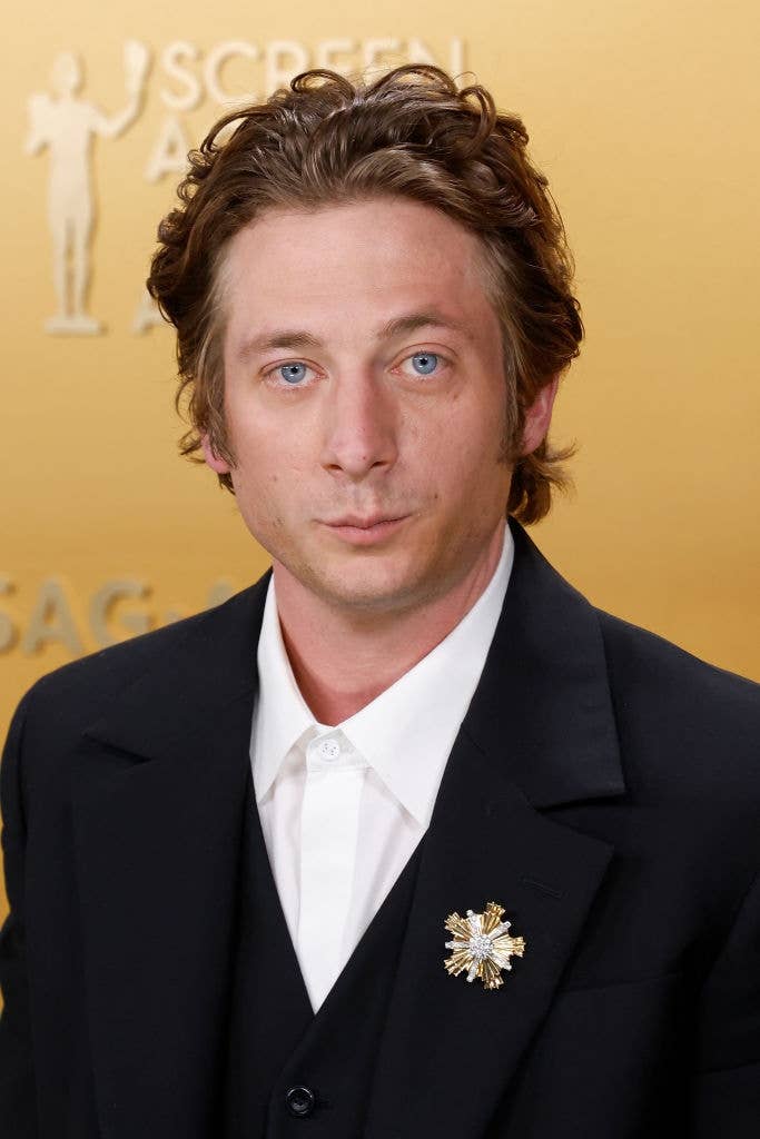 Person in formal black suit with a decorative brooch against a patterned backdrop