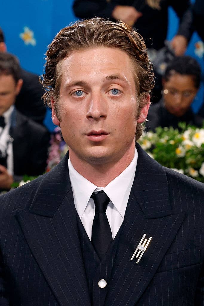Person in a classic pinstripe suit with a unique brooch, at a formal event. People and floral décor are in the background