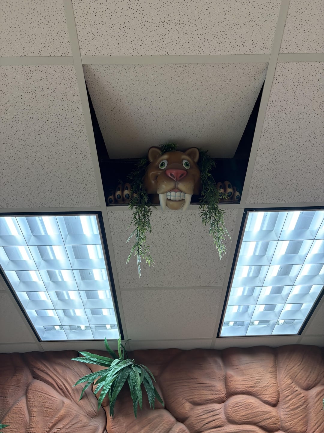 A cartoon animal face with a wide grin peeks through a ceiling tile surrounded by greenery, in an office setting with fluorescent lights