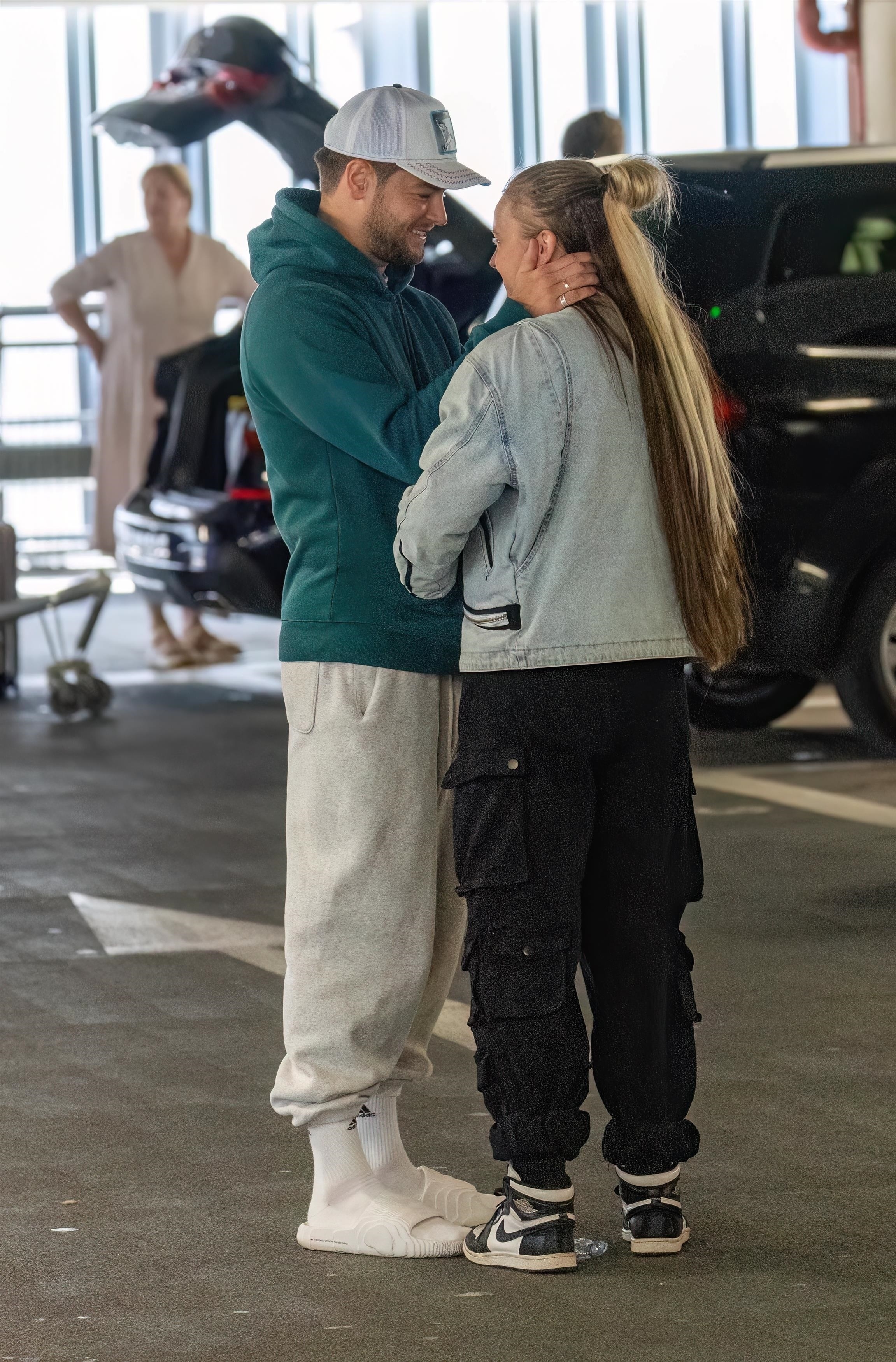Two people stand closely in a parking garage, embracing and smiling at each other. One wears casual sweats, the other a denim jacket and black cargo pants