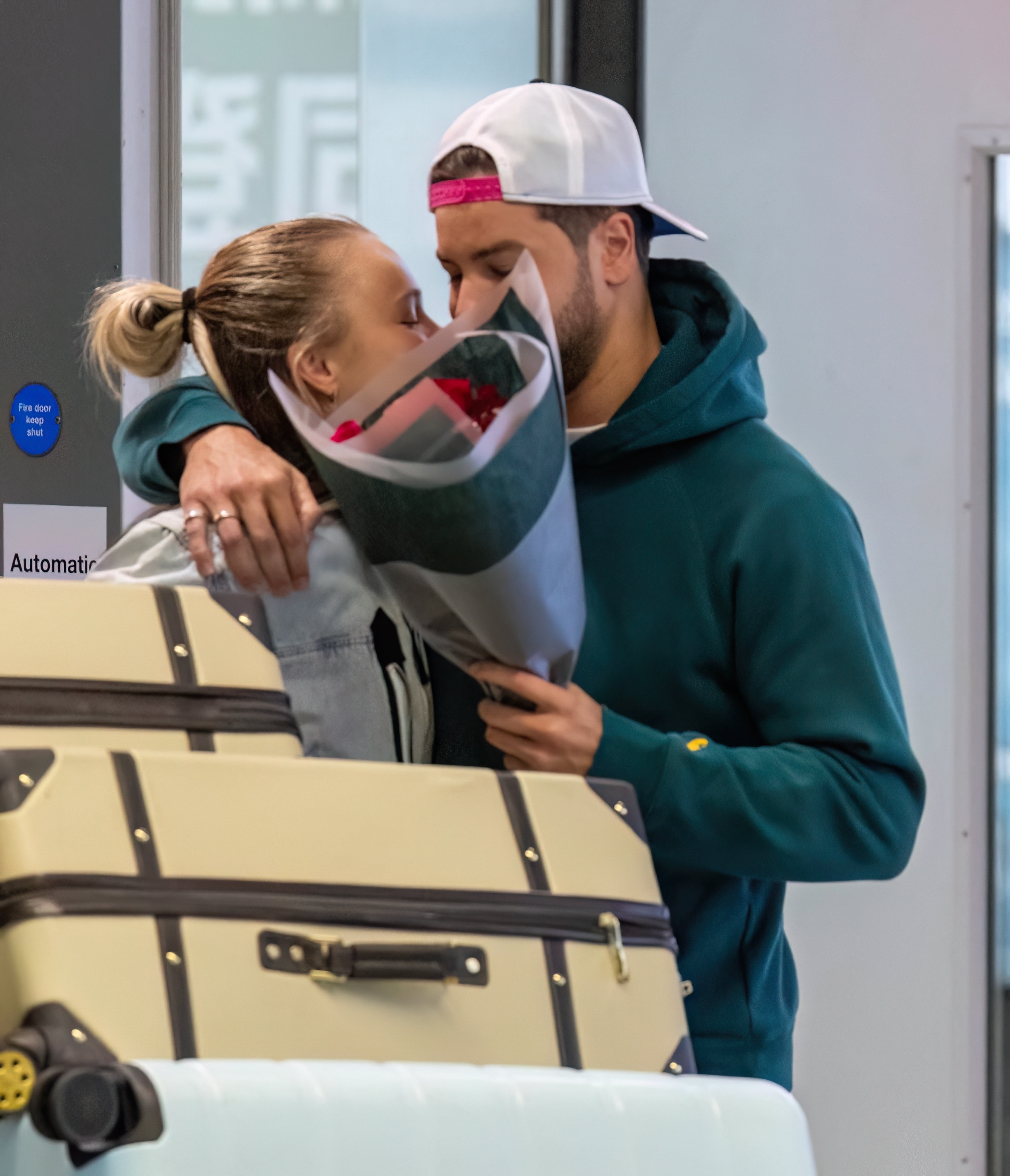 A couple embraces at an airport terminal, holding a bouquet of roses near luggage