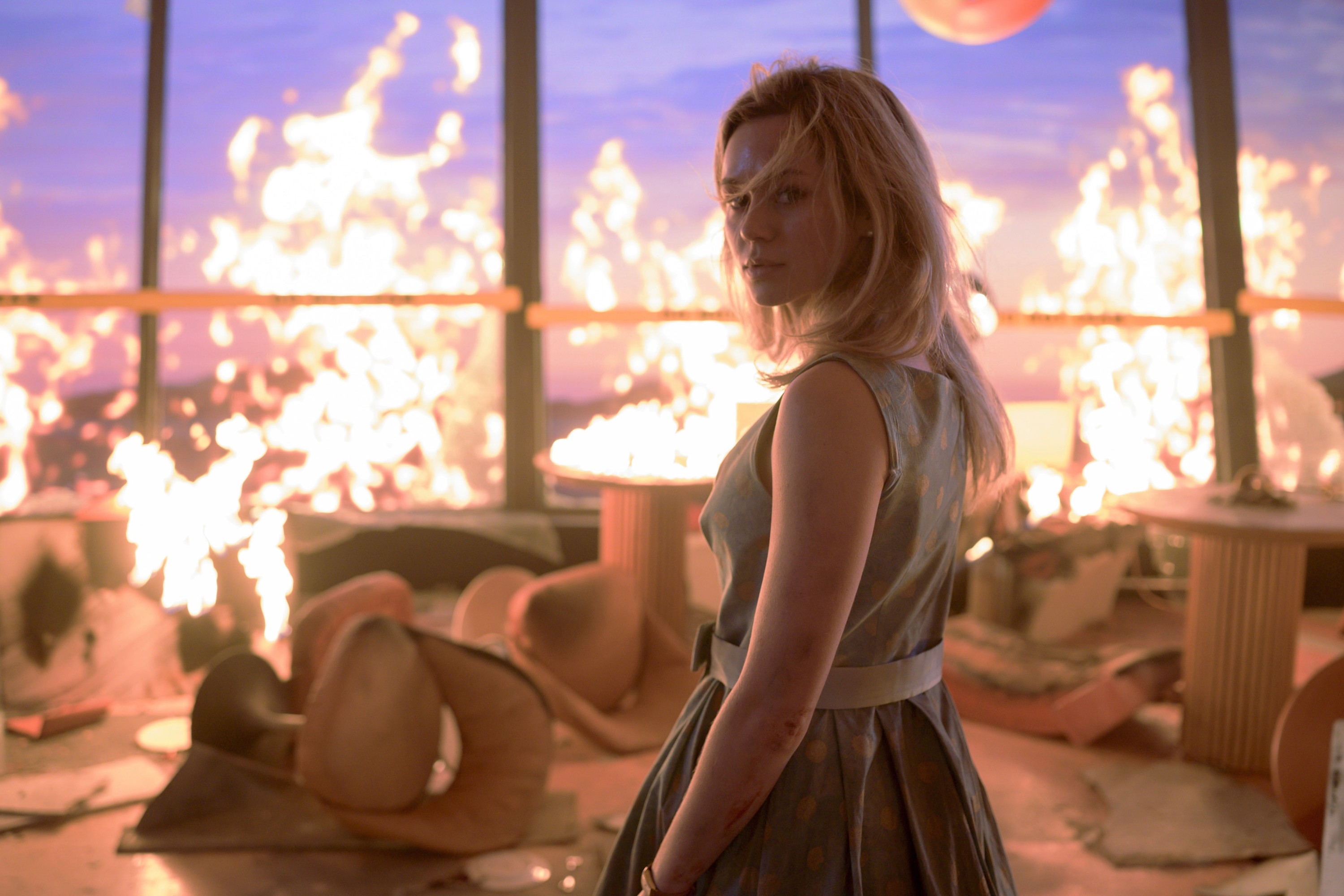 Woman in a stylish dress stands amid burning debris inside a room with large windows