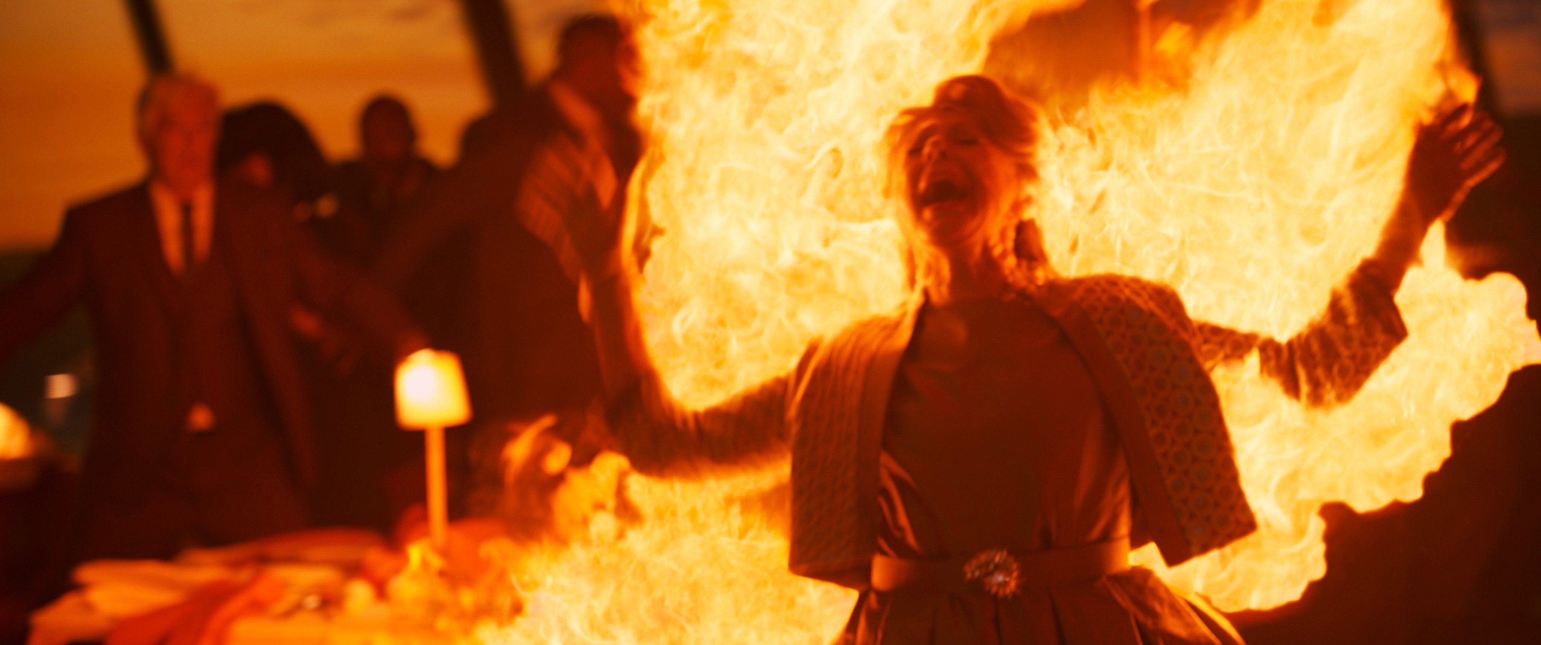 A person stands with arms spread wide, surrounded by flames, in a dramatic scene at a dimly lit gathering