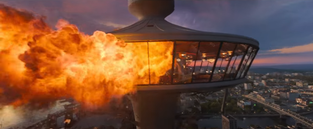 Explosion engulfs a futuristic, glass-walled tower extending outward, with a cityscape in the background