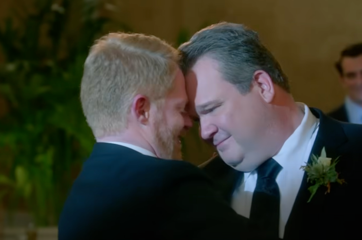 Two men in suits share a joyful intimate moment, foreheads touching, with one wearing a boutonniere. A man in a suit appears in the background