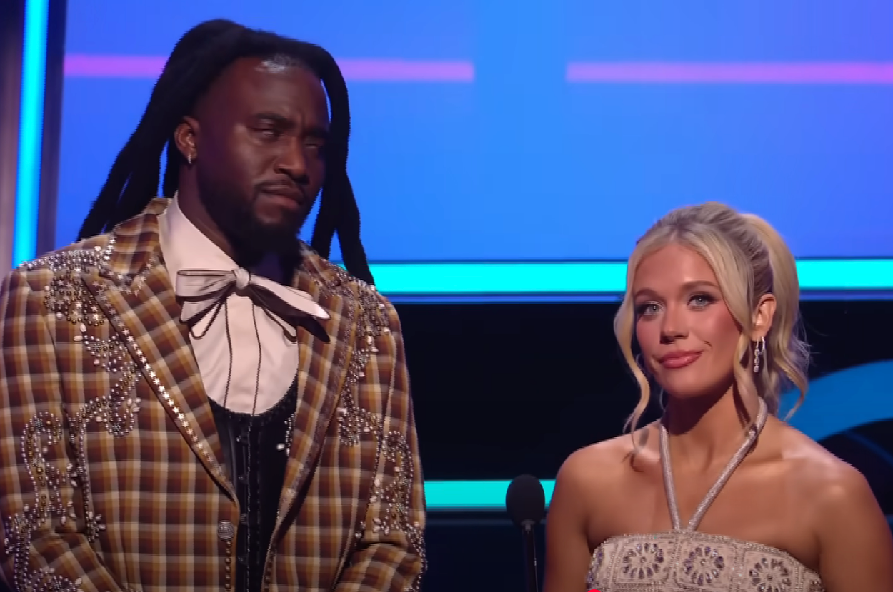 Two people stand onstage. The man wears a plaid suit with embellishments, and the woman wears a strapless dress with a detailed pattern