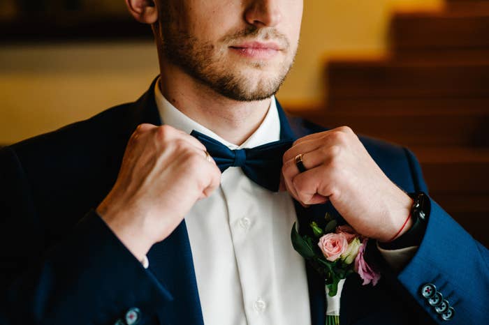 A man in a suit adjusts his bow tie while wearing a boutonniere of small flowers