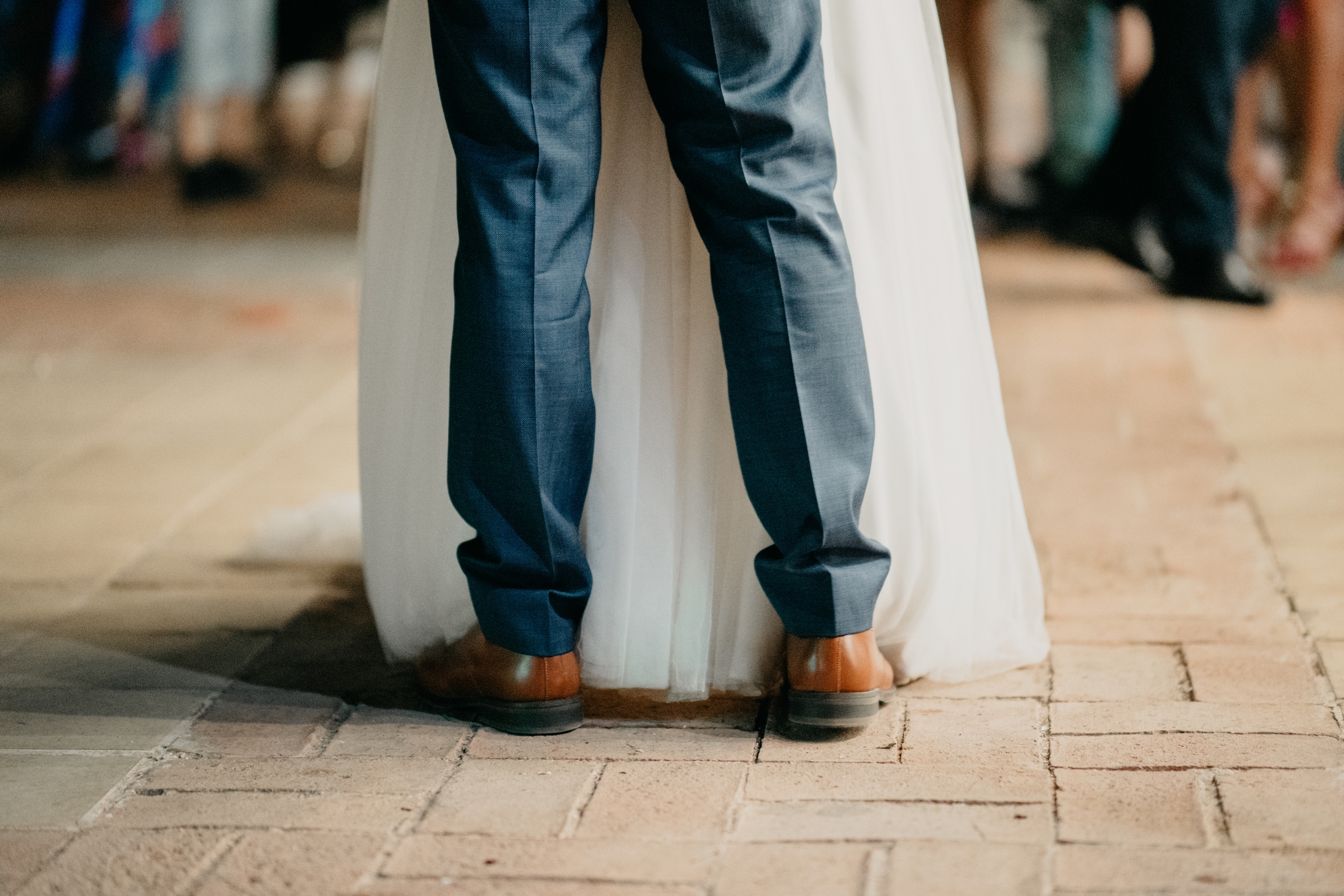 Back view of a couple dancing; person in suit, other in a long gown, surrounded by blurred background of guests