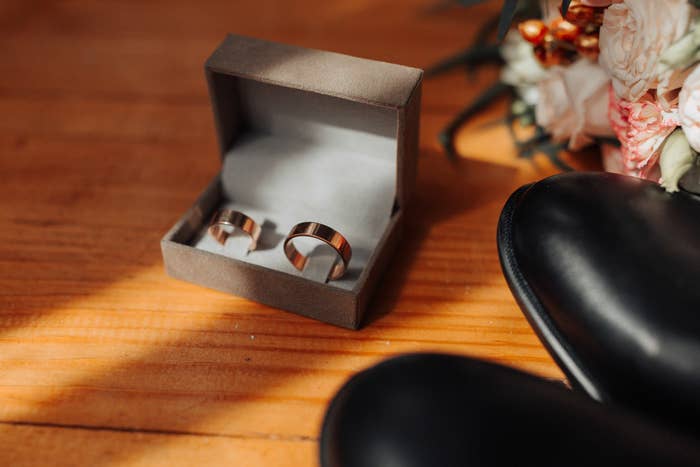 Two gold rings in an open box on a wooden surface, next to a bouquet and black shoes