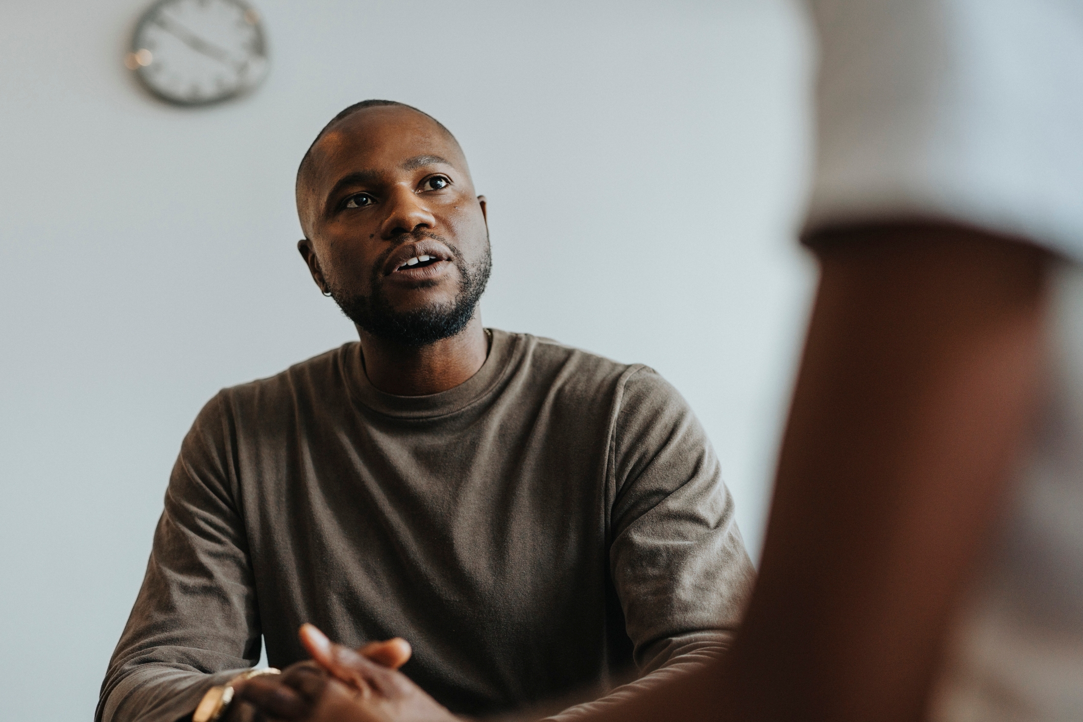 A person attentively listening with hands clasped, wearing a casual long-sleeve shirt in an indoor setting. A clock is visible on the wall