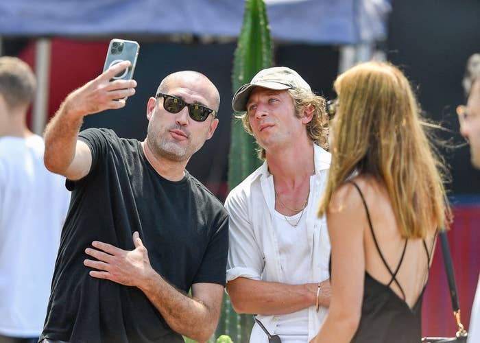 Three people take a selfie at an outdoor event. The man holding the phone wears sunglasses and a T-shirt. Another person wears a casual shirt and cap