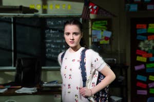 Person with floral-patterned sweater and backpack stands in a classroom with a bulletin board and chalkboard in the background