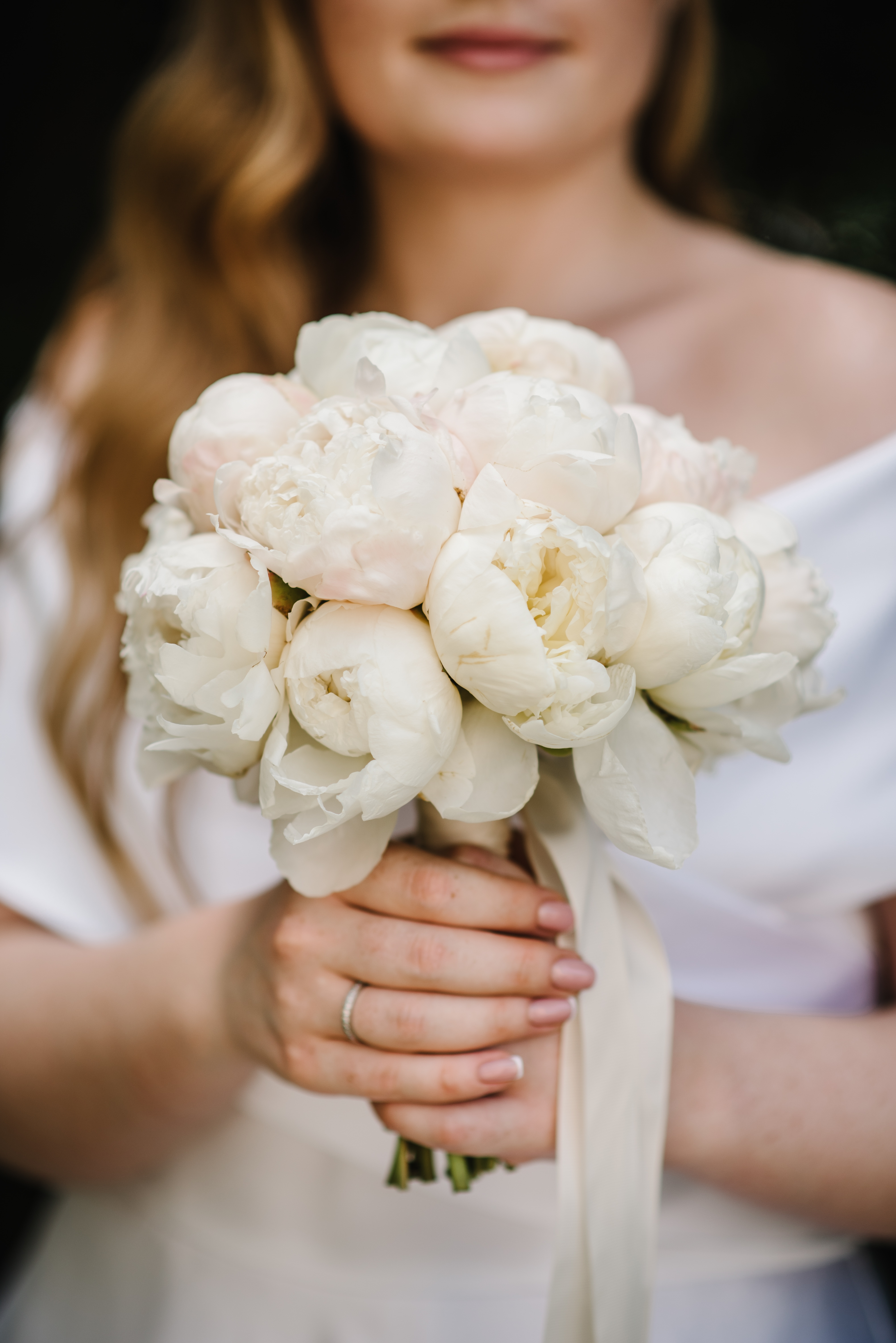Bride holding a bouquet of white peonies in her hands, wearing an off-shoulder dress