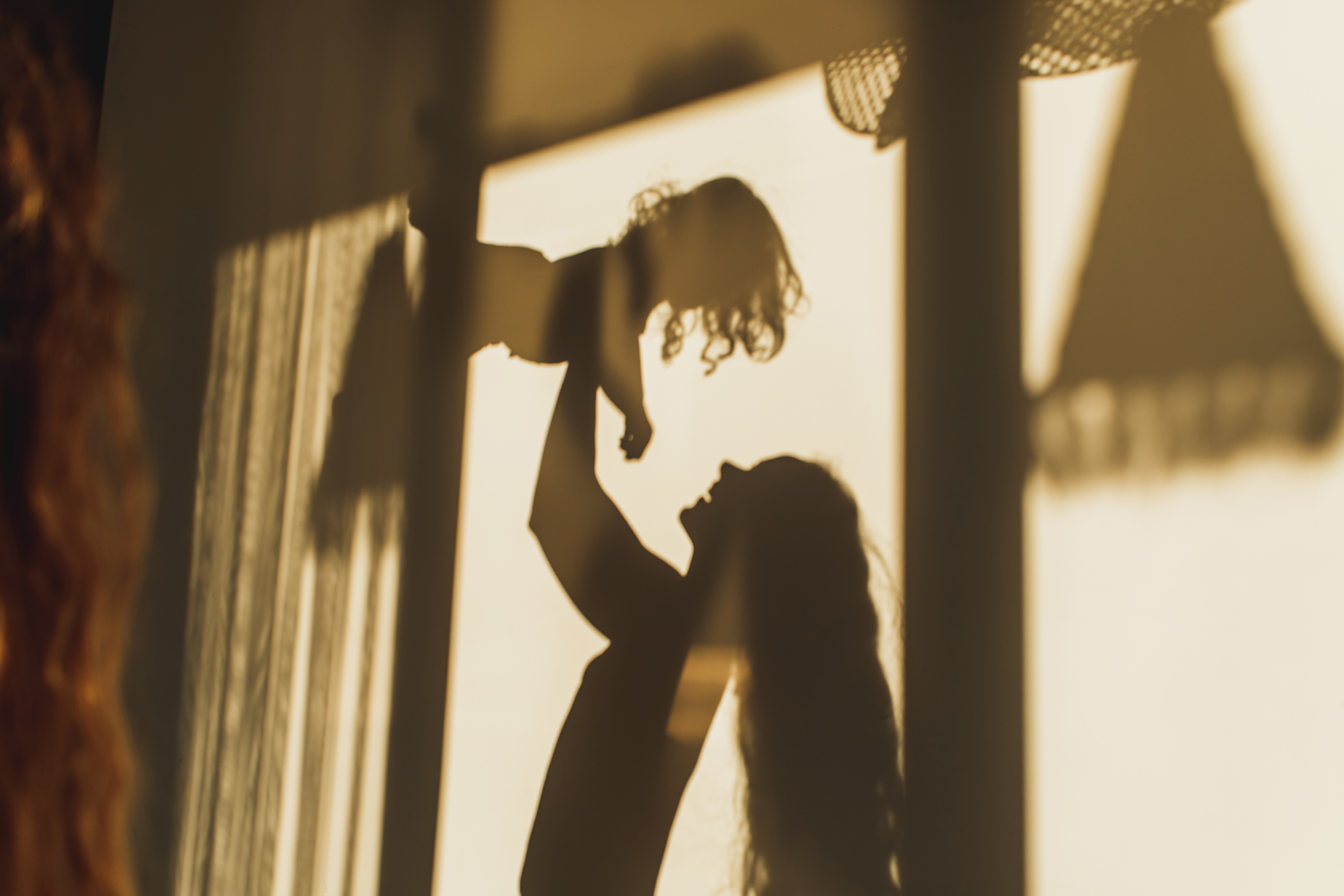 Silhouetted against a curtain, a person joyfully lifts a child in the air, conveying a sense of love and connection